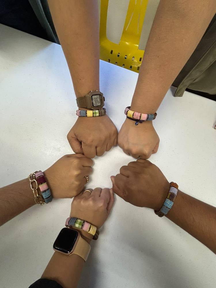Five people joining fists over a table, each wearing a fabric-wrapped values bracelet